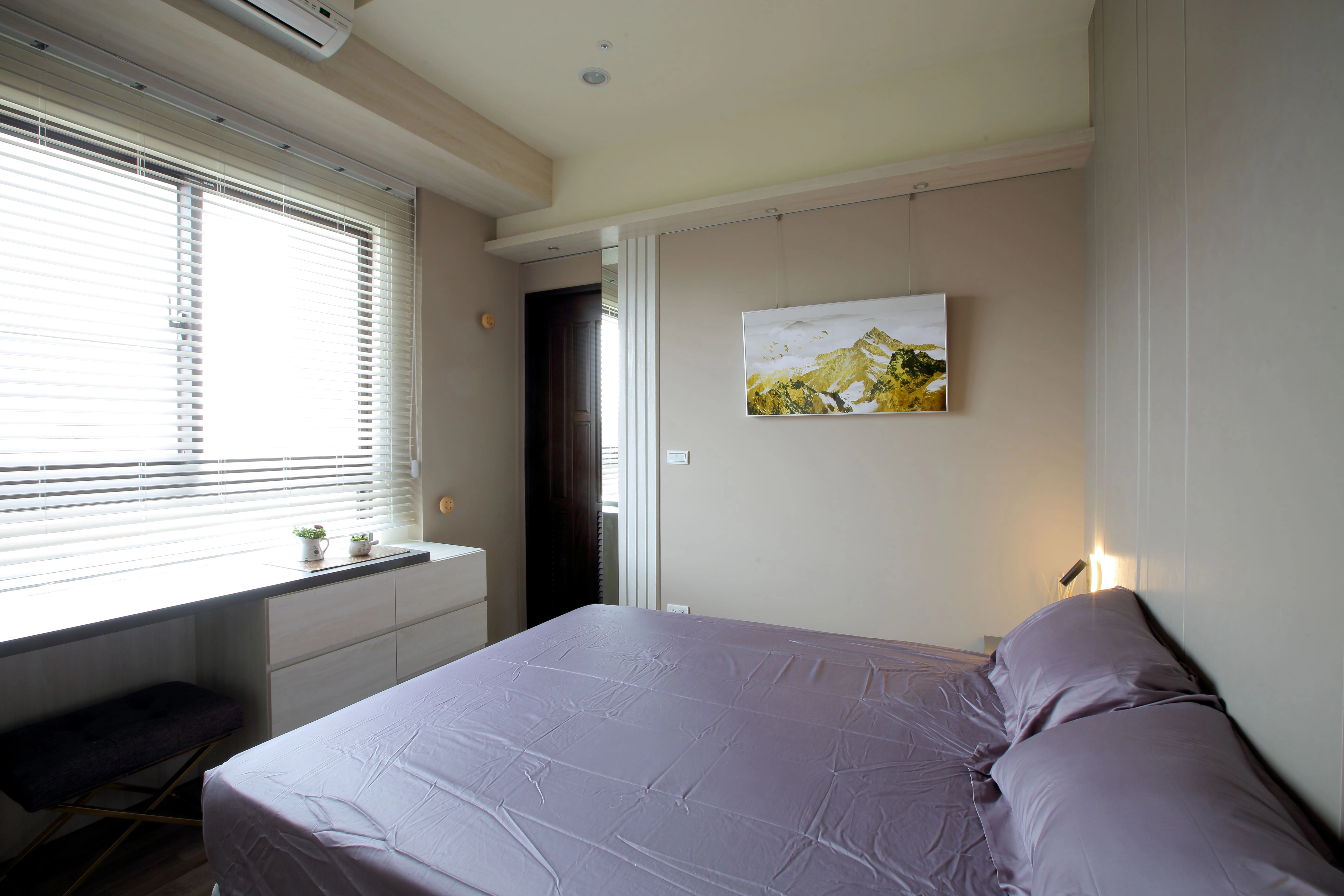 現代簡約臥室設計:暖色調牆面掛畫裝飾,結合系統收納櫃與充足自然光,打造解壓感居家氛圍。